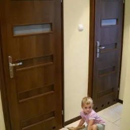 Dwa brązowe drzwi wewnętrzne z przeszkleniami, ułożone na jasnej płytce podłogowej, z małą dziewczynką siedzącą na podłodze i bawiącą się butami.